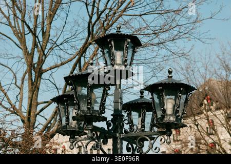 Dekorative Straßenlaterne aus schwarzem Metall mit mehreren Lampen gegen Bäume und Himmel auf dem Balkan, Bosnien. Konzept des Stadtdesigns, historische Straßenbeleuchtung Stockfoto