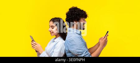 Junges Paar, das Smartphones benutzt, während es sich vor einem hellen gelben Hintergrund Rücken an Rücken lehnt Stockfoto