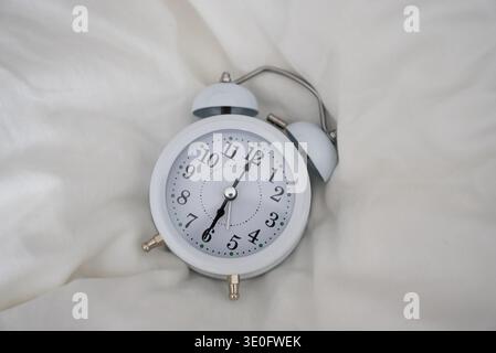 Ein klassischer weißer Wecker mit schwarzen Zahlen und Zeigern auf einer glatten weißen Stoffoberfläche, beleuchtet von weichem natürlichem Licht, das einen Sinn weckt Stockfoto