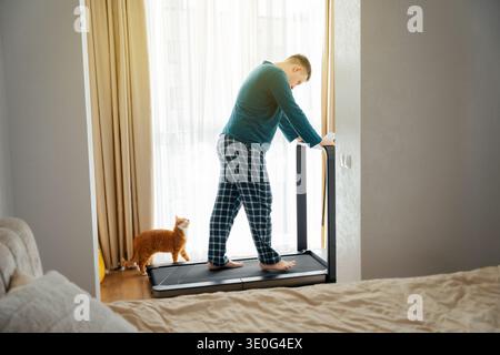 Ein 35-jähriger Kaukasier mit seiner Ingwerkatze läuft an einem sonnigen Tag auf einem Laufband in seiner Wohnung. Heimtraining und Cardio-Training. Stockfoto