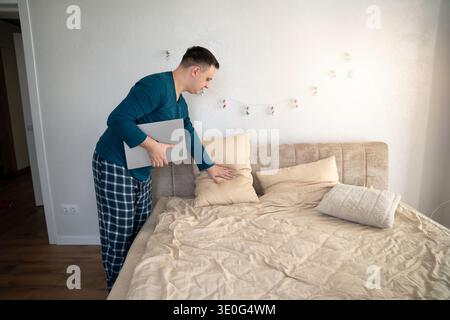 Ein wunderschöner 35-jähriger kaukasier passt sein Kissen an und sitzt mit einem Laptop in seinem Schlafzimmer auf seinem Bett. Fernarbeit, Freizeit und freiberufliches Konzept Stockfoto