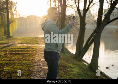 Ein glücklicher junger, schöner, brünette 35-jähriger Mann macht ein Selfie, während er in einem Park in der Nähe eines Sees mit Enten spaziert. Outdoor-Erholung und Lifestyle. Herbst s Stockfoto