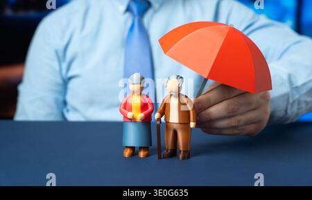 Finanzielle Sicherheit, Lebensversicherung und Seniorenschutz. Der Versicherungsvertreter hält einen roten Schirm über den Figuren eines älteren Paares. Stockfoto