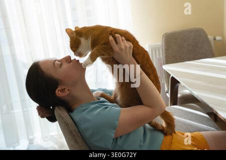Eine brünette kaukasierin küsst und spielt mit einer Ingwerkatze, während sie in einer Küche sitzt. PET Love Konzept. Stockfoto