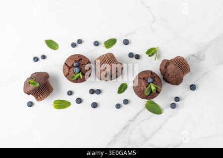 Frisch gebackene Muffins mit Schokoladenchips, Heidelbeeren und Minzblättern auf weißem Marmortischhintergrund. Schokoladen-Cupcake-Nahaufnahme als Nachtisch Stockfoto