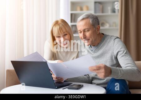 Seniorenpaar, das Dokumente überprüft, während er zu Hause mit Laptop am Tisch sitzt Stockfoto