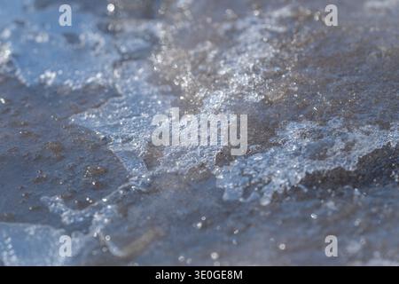 Eis schmilzt schnell. Taufende Eiskristalle schmelzen über feuchtem, dunklem Grund. Der flache Fokus hebt winzige Facetten schmelzender Eiskristalle im Frühling auf dem Boden hervor Stockfoto