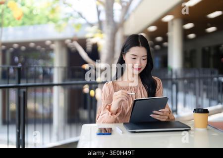 Die junge asiatische Frau konzentriert sich mit einem Stift auf ihr digitales Tablet. Sie studiert oder arbeitet im Freien und genießt moderne Konnektivität. Stockfoto