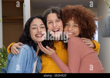 Verschiedene Freundinnen stehen, lehnende Köpfe lächeln beim Eingang mit Senfpullover und Pflanzen. Trio, Begleiter, Wärme, Glück, Foyer, Innenraum, Stockfoto