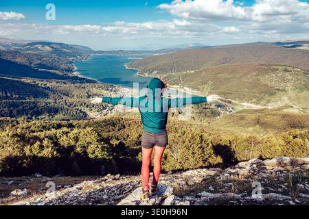 Blick auf eine Figur in einer blauen Jacke und roten Leggings, die die weite Landschaft umgibt, mit einem glitzernden See inmitten sanfter Hügel, Mavrovë, North Mac Stockfoto