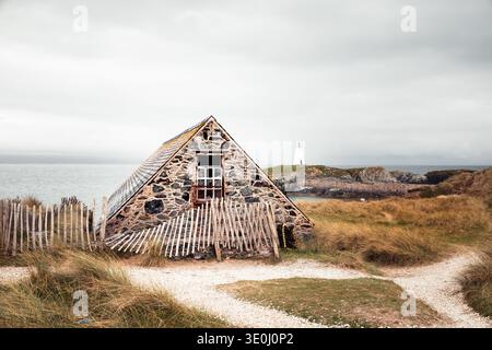 Eine Ruine aus Stein mit eingestürzten Holzzäunen auf Llanddwyn Island, mit dem Leuchtturm von Tŵr Mawr auf der Landzunge hinter Anglesey. Stockfoto