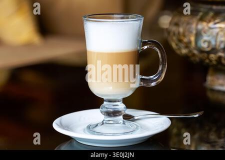 Lagen Latte Macchiato mit Milchschaum, serviert in transparentem Glas auf reflektierendem Kaffeetisch. Elegante Präsentation, geeignet für Gastfreundschaft und Restaurant Stockfoto