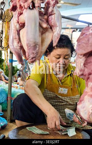 Eine Frau zählt Geld auf einem Schneidebrett auf einem kambodschanischen Fleischmarkt nach einem Verkauf. Rohes Fleisch hängt um sie herum, während sie einen Verkauf macht. Stockfoto