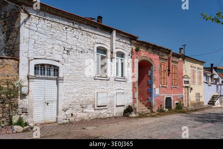Alte Häuser säumen eine Kopfsteinpflasterstraße in einem Dorf. Einige Häuser haben farbenfrohe Wände und viele zeigen Alterspuren. Die Sonne scheint hell darüber und hebt die einfachen Strukturen hervor. Stockfoto