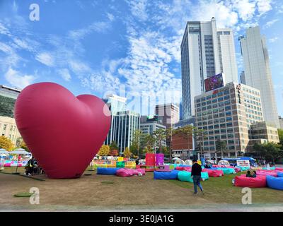 Besucher versammeln sich um eine große Herzskulptur, während sie einen sonnigen Tag in einem Park genießen, der von hohen Gebäuden in Seoul umgeben ist. Stockfoto