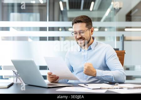 Geschäftsmann im modernen Büro feiert gute Nachrichten, hält ein Dokument und ballt seine Faust mit einem fröhlichen, triumphierenden Lächeln an seinem Schreibtisch, einem Laptop in der Nähe, in professioneller Kleidung Stockfoto
