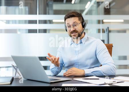 Ein Geschäftsmann mit Headset und Brille ist Videokonferenzen auf seinem Laptop, der aktiv Gesten gibt, während er während eines virtuellen Meetings in einer modernen Büroumgebung kommuniziert Stockfoto