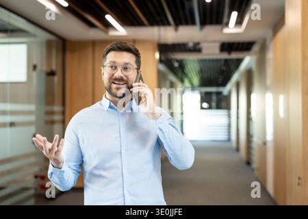 Lächelnder professioneller Mann mit Brille, der sich auf einem Mobiltelefon unterhalten kann, während er in einem modernen Korridor des Firmengebäudes steht und über Geschäfte spricht Stockfoto