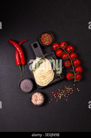 Scheiben von festem Käse mit Kräutern, roten Chilischoten, Kirschtomaten auf der Rebe und verschiedenen Gewürzen in Schüsseln angeordnet und auf dunkler Textur verstreut Stockfoto
