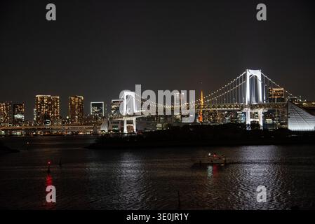Die Regenbogenbrücke überspannt die Bucht von Tokio unter einem dunklen Himmel mit dem dahinter leuchtenden Tokyo Tower, einem vorbeifahrenden Boot und der gesamten Skyline der Stadt dahinter. Stockfoto
