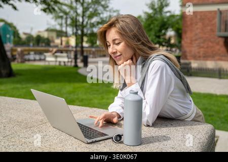 Lächelnde, glückliche Geschäftsfrau arbeitet am Sommertag gern im Freien an einem Laptop, was die Vorteile der Fernarbeit ist Stockfoto