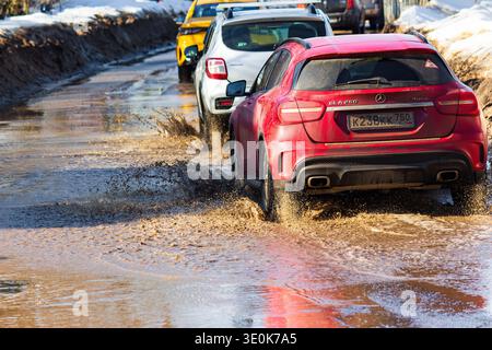 Der rote Mercedes GLA 250 SUV plätschert kräftig durch tiefe, schlammige Frühlingspfützen auf unebener Straße. Schwierige russische Straßenbedingungen nach Schneeschmelze: R Stockfoto