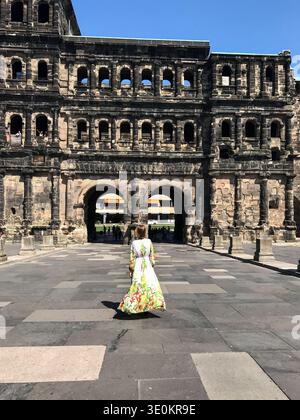 Eine nicht erkennbare Frau, die von hinten gesehen wird und ein langes, fließendes Sommerkleid mit Blumenmuster trägt, geht in Richtung der historischen Porta Nigra. Römische Architektur Stockfoto
