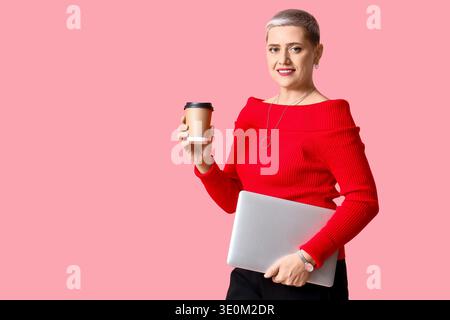 Glückliche reife blonde Frau mit kurzen Haaren, Laptop und Tasse Kaffee auf rosa Hintergrund Stockfoto