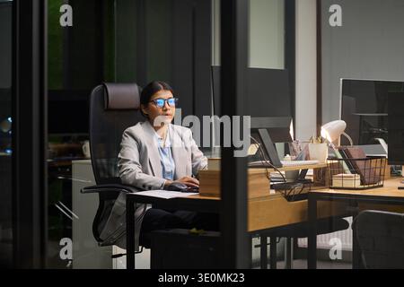 Junge Frau, die nachts am Schreibtisch sitzt und im modernen Büro am Computer arbeitet, Brille und Bürokleidung trägt, sich auf den Bildschirm konzentriert und von Bürogeräten umgeben ist Stockfoto