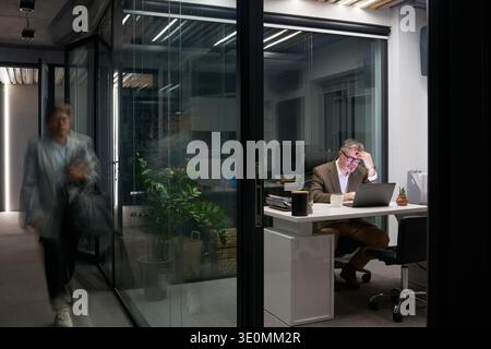 Kaukasischer Mann mittleren Alters, der nachts am Schreibtisch sitzt und in einem modernen Büro an einem Laptop arbeitet, mit der Hand den Kopf hielt und gestresst aussah, während ein unscharfer junger Erwachsener an einer Glaswand vorbeiging Stockfoto