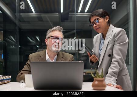 Kaukasischer Mann mittleren Alters sitzt am Schreibtisch mit Laptop, während die junge hispanische Frau neben ihm mit einem Smartphone über die Geschäftsstrategie in einem modernen Büroumfeld diskutiert Stockfoto