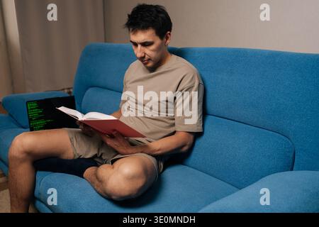 Erfahrene Entwickler, die auf blauem Sofa sitzen, studieren das Lehrbuch, während der Laptop grüne Codelinien zeigt, die fokussiertes Fernlernen und Kompetenzentwicklung veranschaulichen Stockfoto