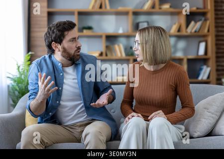 Das junge Paar hat ein ernsthaftes Argument und Gesten, während es auf einem Sofa in seinem Wohnzimmer sitzt, was auf Beziehungsprobleme und Kommunikationsstörungen hinweist Stockfoto