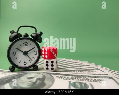 Kleiner schwarzer Wecker und zwei gestapelte Würfel auf einem Fan von US-Dollar-Scheinen auf grünem Hintergrund. Stockfoto