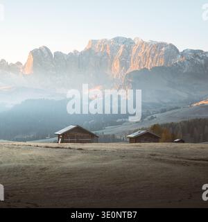 Blick auf rustikale Holzchalets in einem frostigen Feld unter den majestätischen, sonnengeküssten Dolomiten, die in goldenes Licht und ätherischen Nebel getaucht sind, Alpe di Stockfoto