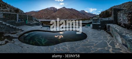 Ein breiter Panoramablick zeigt die Steinarchitektur der heißen Quellen von Panamik gegen die riesigen Grate des Karakorum. Stockfoto