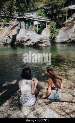 Zwei Freunde sitzen an einem felsigen Fluss und genießen Getränke in einer sonnigen Umgebung im Freien Stockfoto
