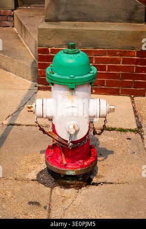 Ein alter Feuerhydrant mit abblätternder Farbe befindet sich auf einem Bürgersteig aus Beton, der an einer roten Ziegelwand positioniert ist. Stockfoto
