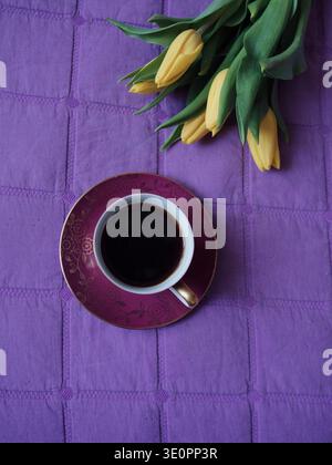 Gelber Tulpenstrauß und Vintage Coffee Cup auf lila Decke – flache gemütliche Morgenszene Stockfoto