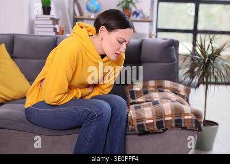 Frau in gelbem Hoodie sitzt auf der Couch und hält ihren Magen an einem ruhigen Nachmittag zu Hause unangenehm Stockfoto