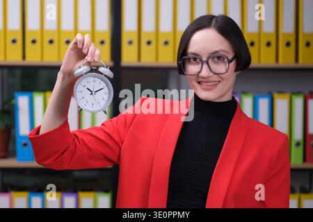 Eine selbstbewusste Frau in einem auffälligen roten Blazer hält einen Wecker und symbolisiert die Bedeutung des Zeitmanagements in einer lebendigen Büroumgebung Stockfoto