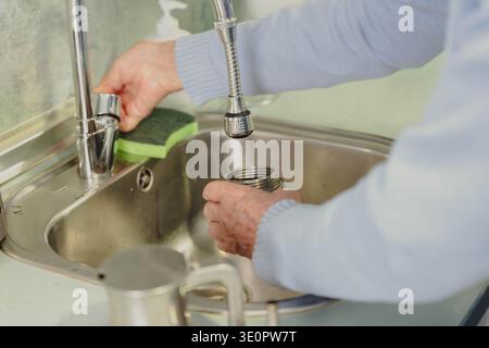 Hände eines älteren Mannes waschen eine Metallkaffeemaschine und einen Schwamm in einem Küchenspüle Stockfoto