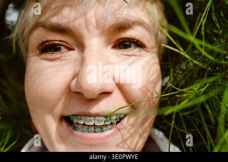 Porträt einer glücklichen Frau, die mit kieferorthopädischen Zahnspangen lächelt und die Natur genießt. Junge Erwachsene Frau mit Zahnspangen draußen zwischen Bäumen und Blumen Stockfoto