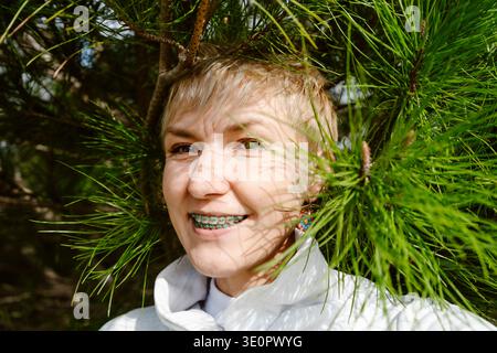 Porträt einer glücklichen Frau, die mit kieferorthopädischen Zahnspangen lächelt und die Natur genießt. Junge Erwachsene Frau mit Zahnspangen draußen zwischen Bäumen und Blumen Stockfoto