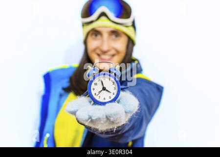 Close up schöne Frau halten Wecker Show in Händen tragen Skifahrer Outfit bereit für draußen isolierte Mock up blaue Studio Hintergrund. Wochenendausflug t Stockfoto