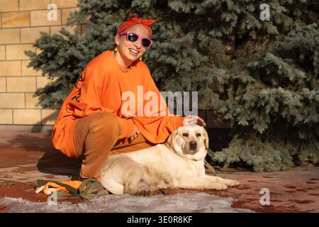 Frau in orangefarbener Kleidung, die neben dem Labrador-Hund sitzt, während der Spaziergang im Freien auf dem Backsteinparkweg läuft. Heller Retriever liegt an der Leine nahe Everg Stockfoto