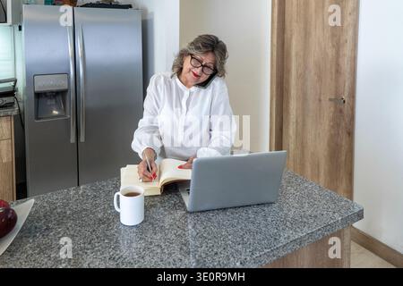 Seniorin jongliert mit Laptop, Telefonat, Notebook und Kaffee und verwaltet ihre freiberufliche Arbeit Stockfoto