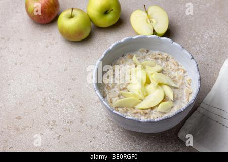Haferbrei mit Milch und frischen Apfelscheiben in einer Keramikschale. Nahrhaftes Frühstück mit Hafer und Obst auf hellem Küchentisch Stockfoto