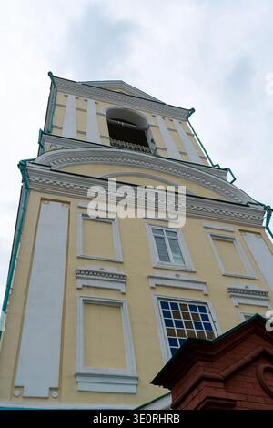 Ein gelb-weißer Kirchturm, der aus einem niedrigen Winkel erfasst wurde und neoklassizistische Details, Säulen und bogenförmige Öffnungen vor einem weichen, bewölkten Himmel zeigt Stockfoto