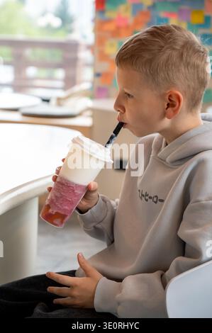 Seitenansicht eines Kindes in einem grauen Hoodie, das ein kaltes Getränk in einem Café genießt. Er hält einen hohen Plastikbecher mit einem bunten Fruchtgetränk und Schaum. Hintergrund Stockfoto